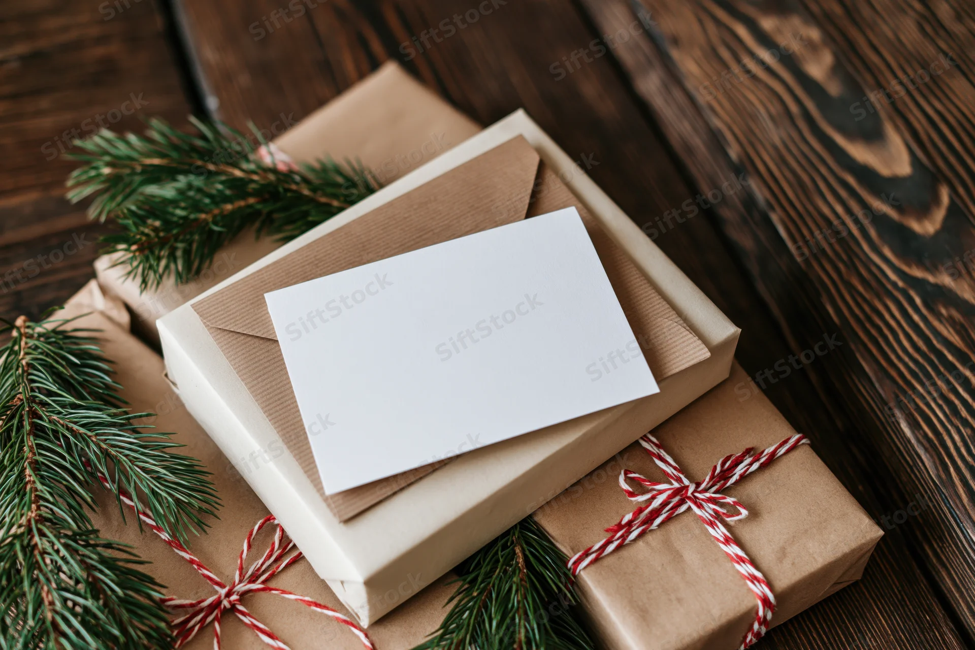 Blank white greeting card on kraft paper envelope with wrapped gifts and pine branches on wooden table