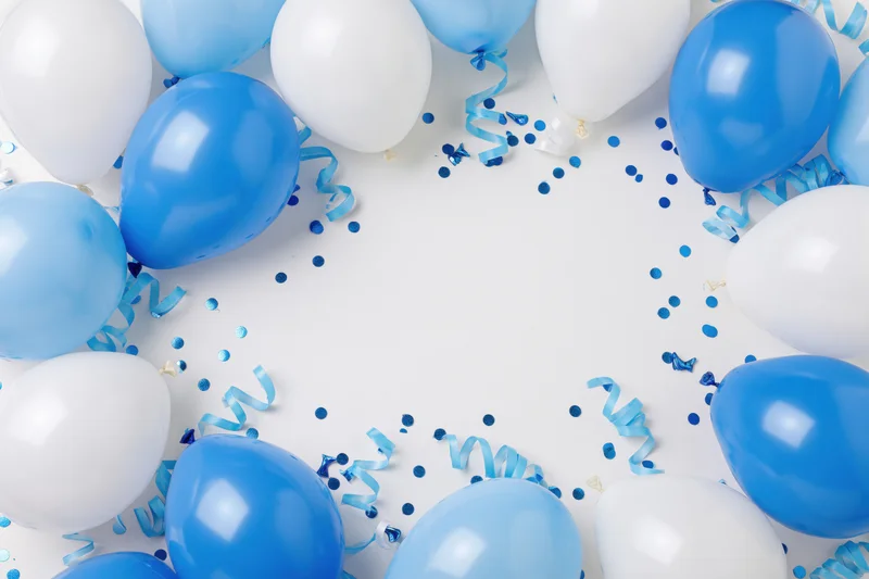 Blue and white balloons arranged in a circular border with confetti and ribbons on a white surface, leaving central copyspace