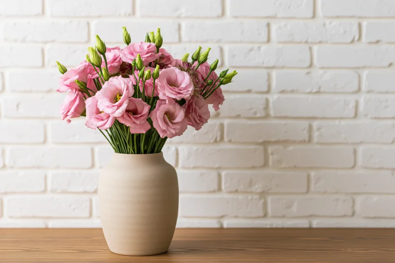 Pink lisianthus bouquet in ceramic vase on wooden table against white brick wall