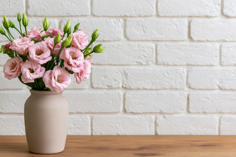 Pink lisianthus bouquet in ceramic vase on wooden table against white brick wall