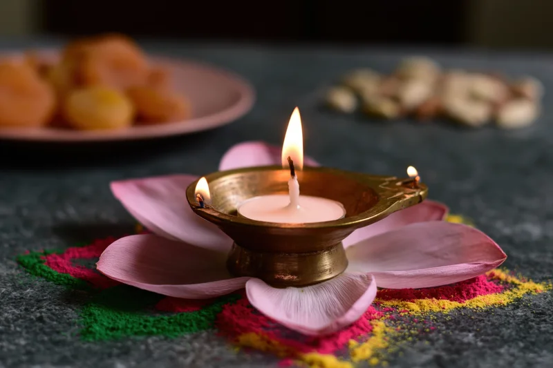 Brass diya with lit candle placed on pink flower petals atop colorful rangoli on dark surface