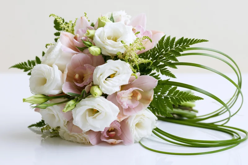 Bridal bouquet with white roses and pink orchids arranged with green ferns and stems on a white surface