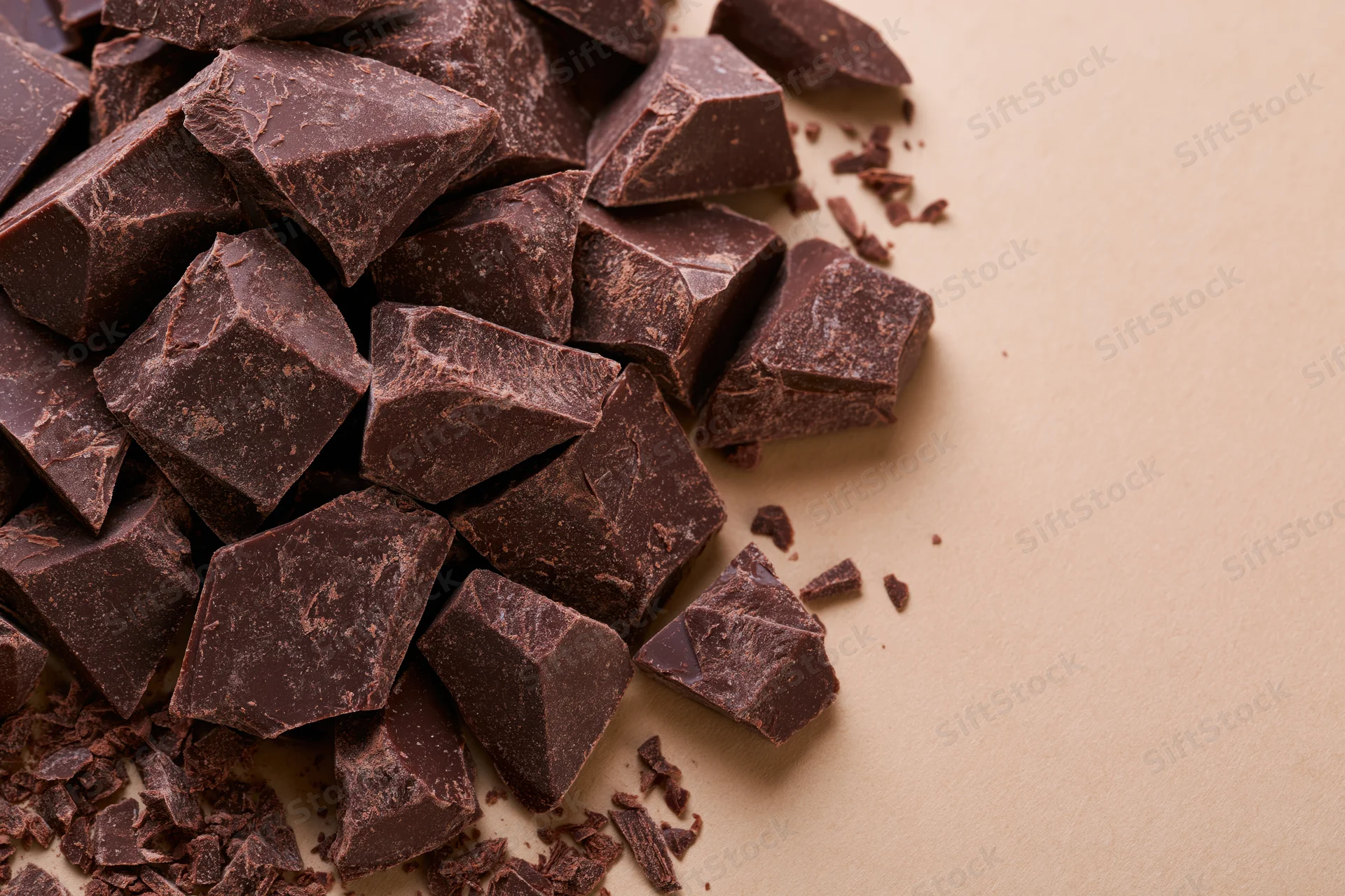 Broken dark chocolate chunks piled loosely on a beige surface with copyspace