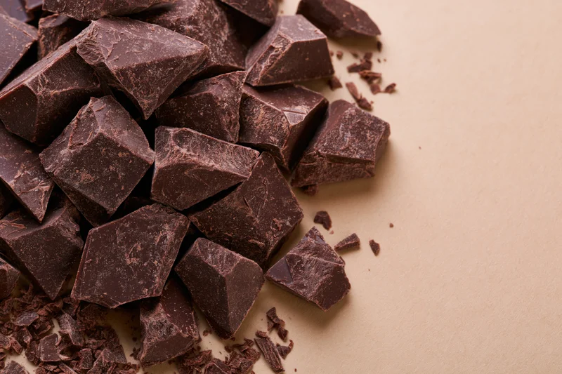 Broken dark chocolate chunks piled loosely on a beige surface with copyspace