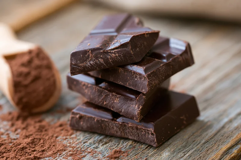 Broken dark chocolate chunks stacked on a rustic wooden surface with cocoa powder nearby