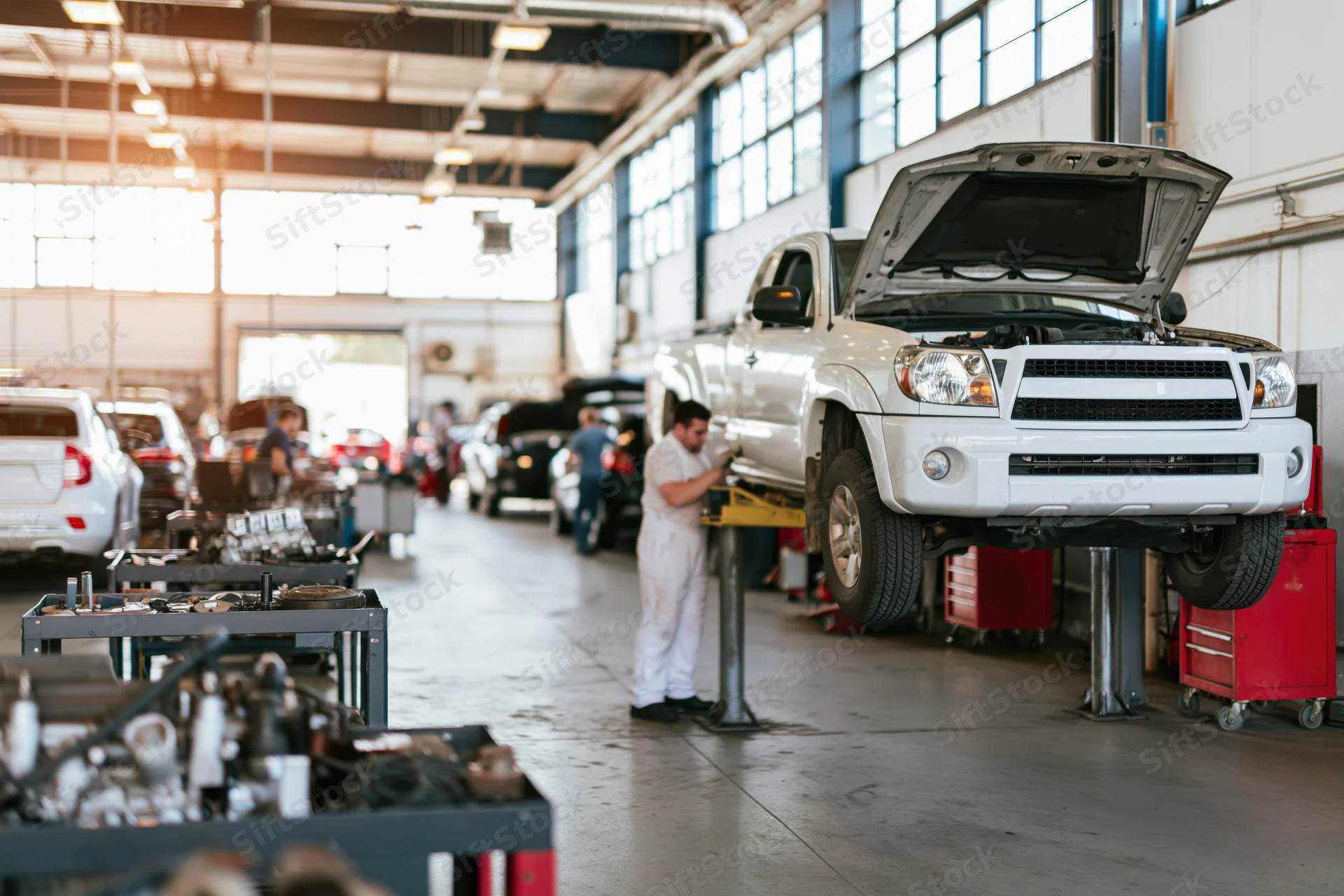 Mechanic working under hood of white pickup truck elevated on lift in automotive repair shop