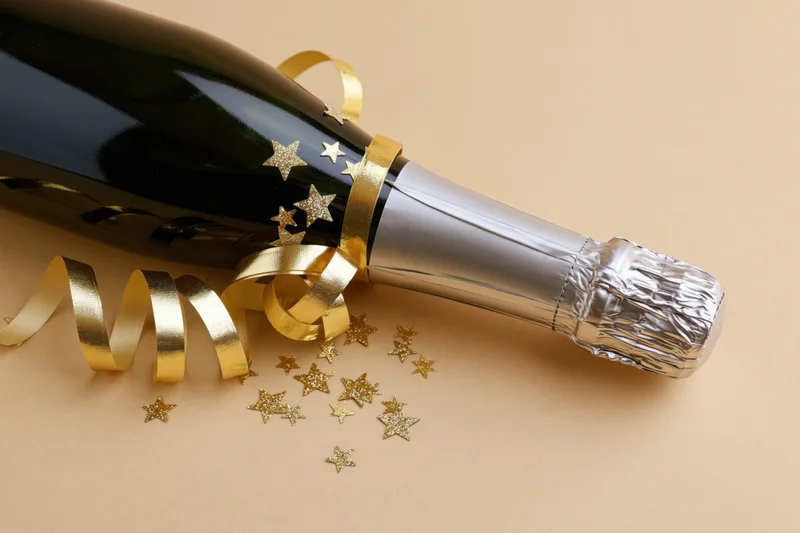 Champagne bottle decorated with gold ribbon and star confetti lying on beige surface