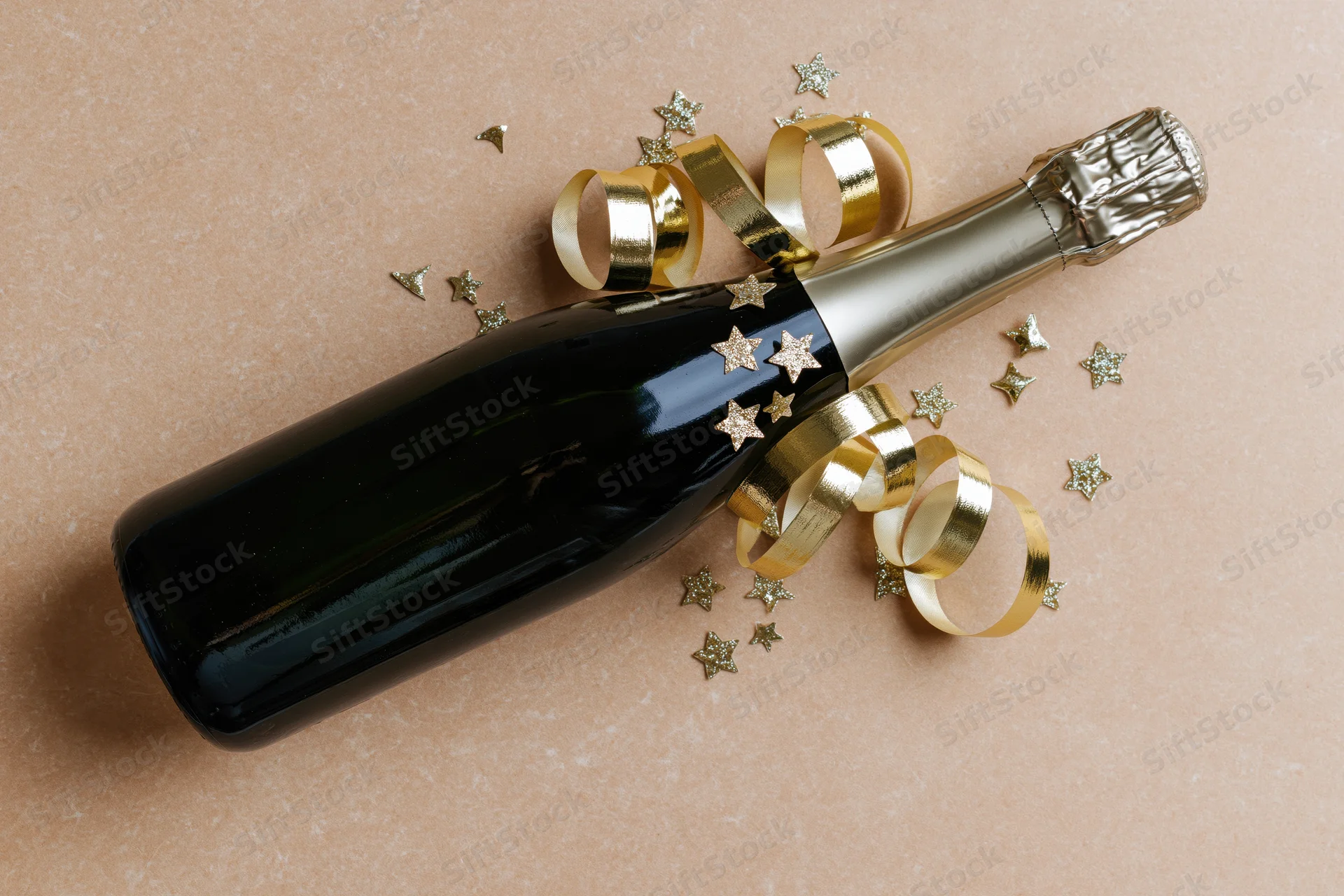 Champagne bottle lying on its side with gold ribbons and star confetti on a pink surface