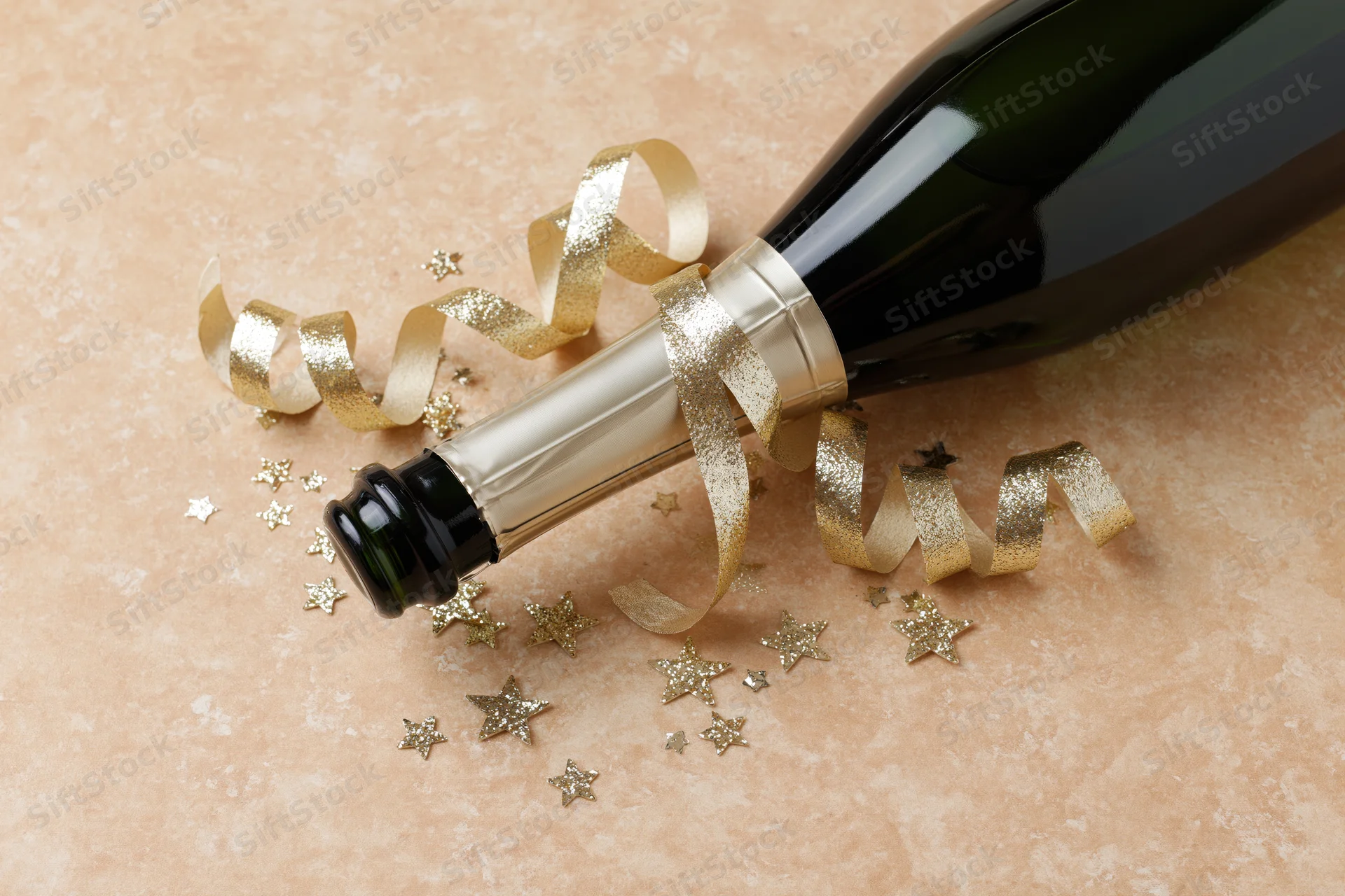 Champagne bottle lying on its side with gold ribbon and star confetti on textured surface