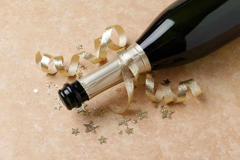Champagne bottle lying on its side with gold ribbon and star confetti on textured surface