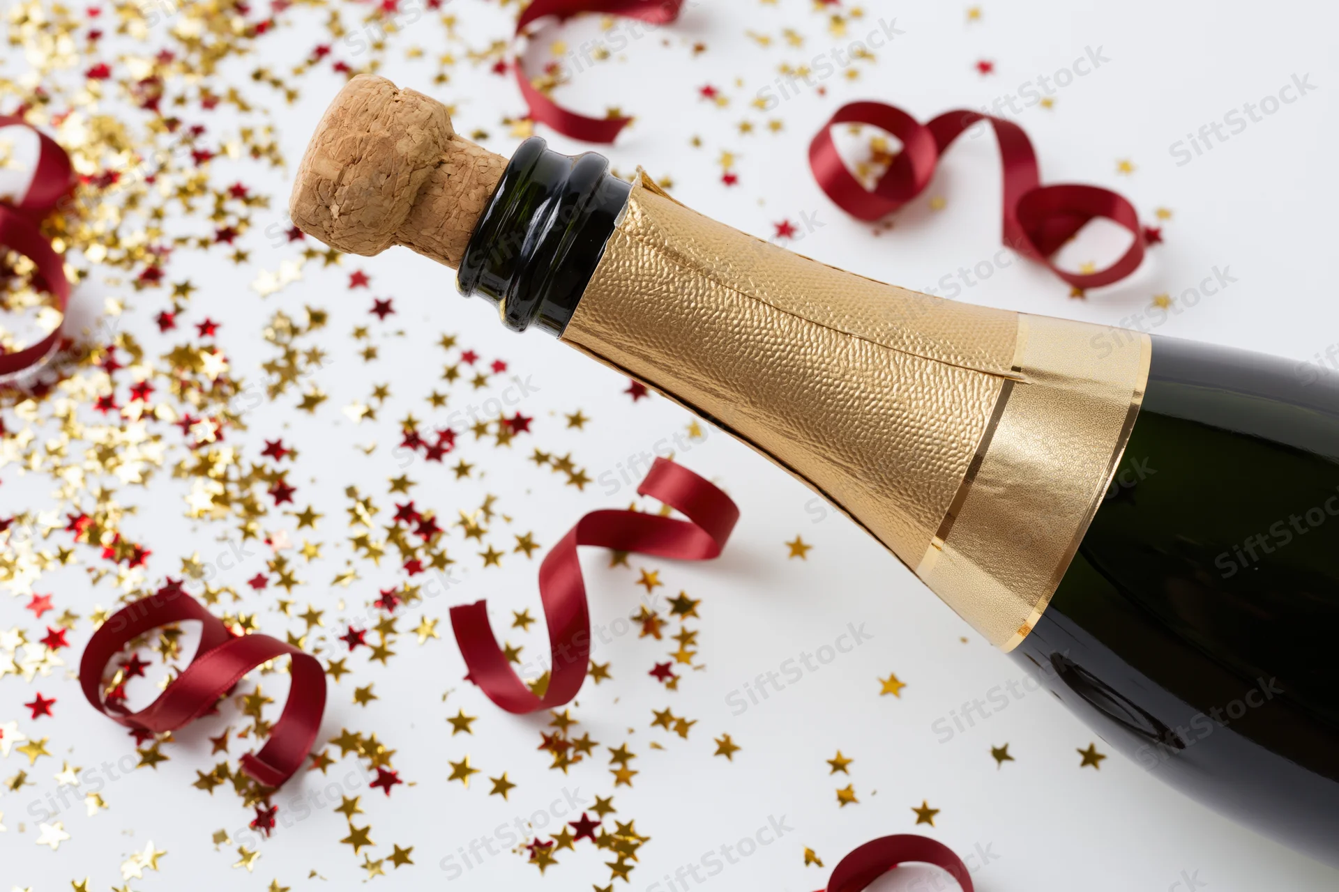 Champagne bottle with cork released lying on white surface surrounded by gold star confetti and red ribbons