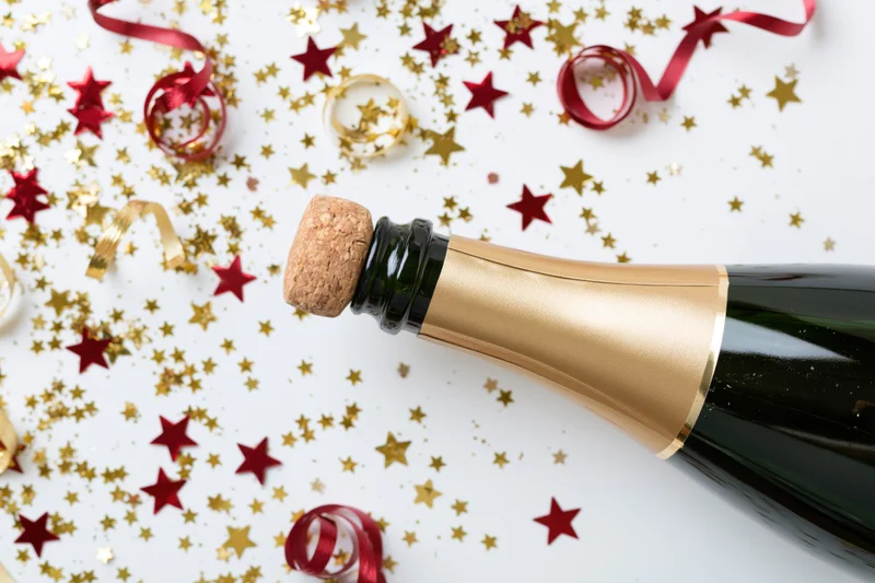 Champagne bottle with cork just released on a white surface scattered with red and gold star confetti and ribbons