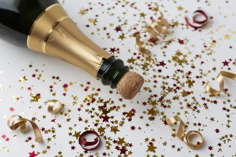 Champagne bottle with cork released lying on white surface surrounded by gold and red star confetti and curled streamers