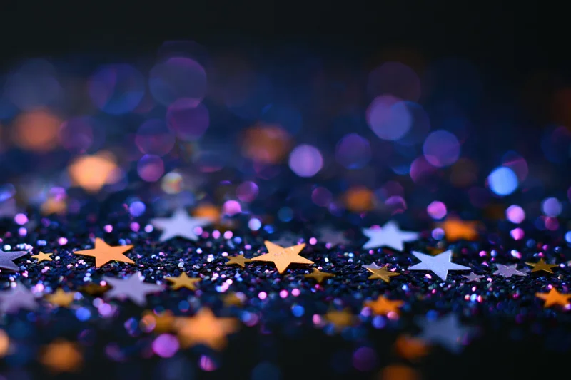 Iridescent glitter and star-shaped confetti scattered on a dark surface with bokeh lights in the background