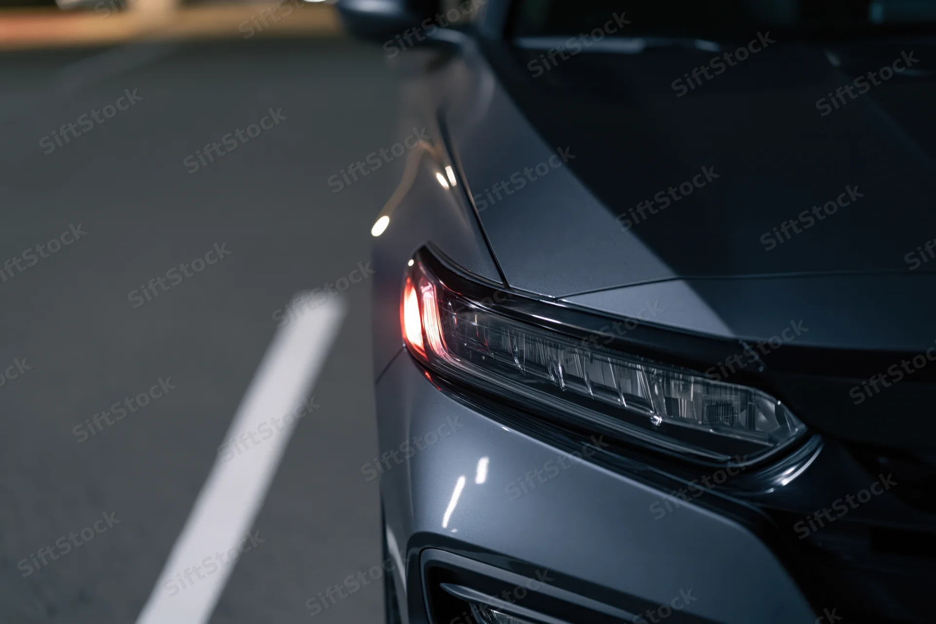Close-up of a dark grey sedan's front headlight and hood corner on a road at night