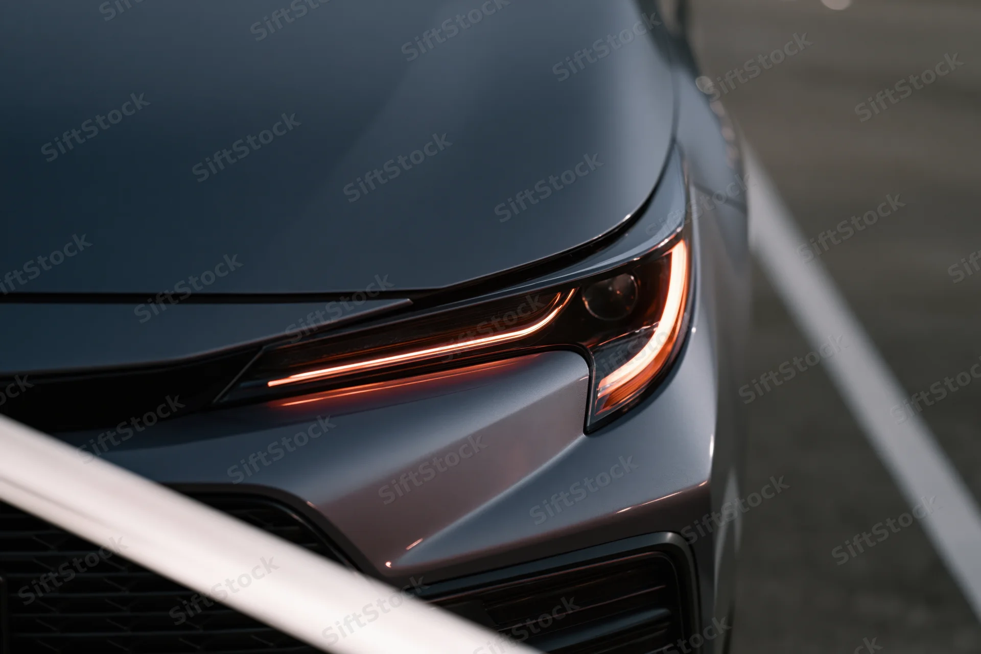 Close-up of a dark grey sedan's front headlight and hood corner on a paved surface with white parking lines