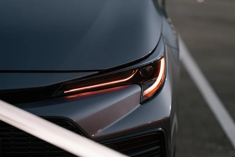 Close-up of a dark grey sedan's front headlight and hood corner on a paved surface with white parking lines