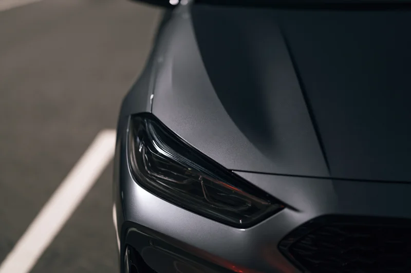 Close-up of a dark grey sedan's front headlight and hood corner on a paved road with a white line