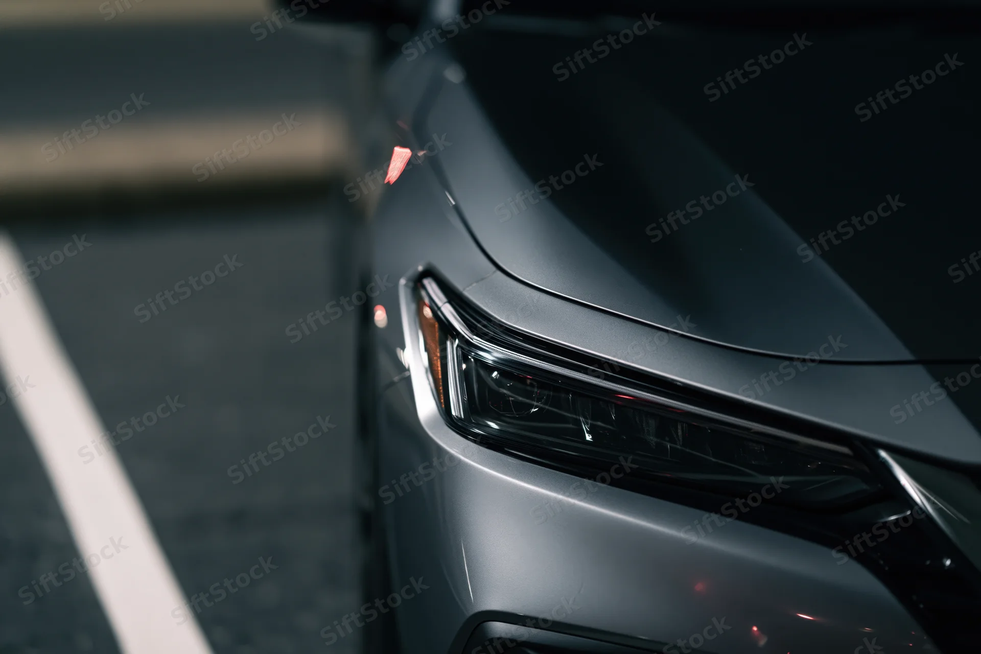 Close-up of a dark grey sedan's front headlight and hood corner on a paved road