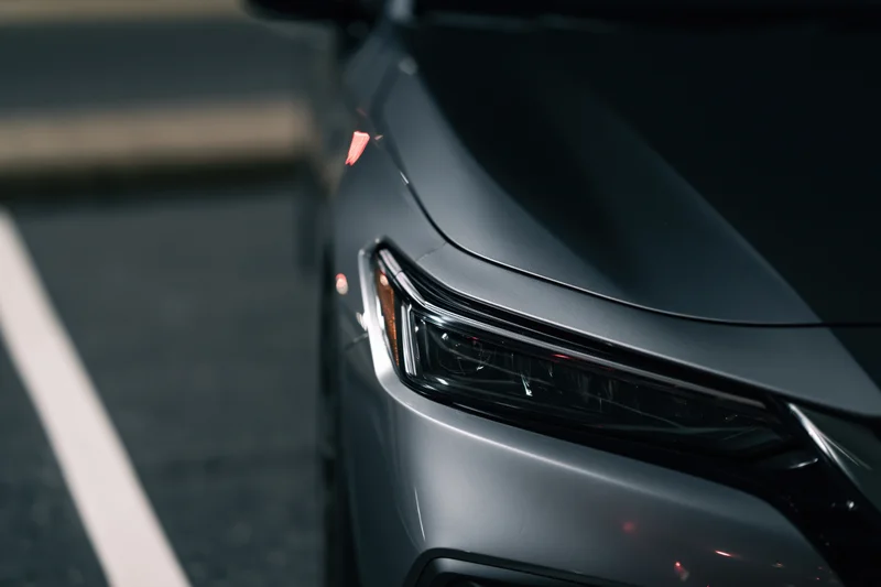 Close-up of a dark grey sedan's front headlight and hood corner on a paved road