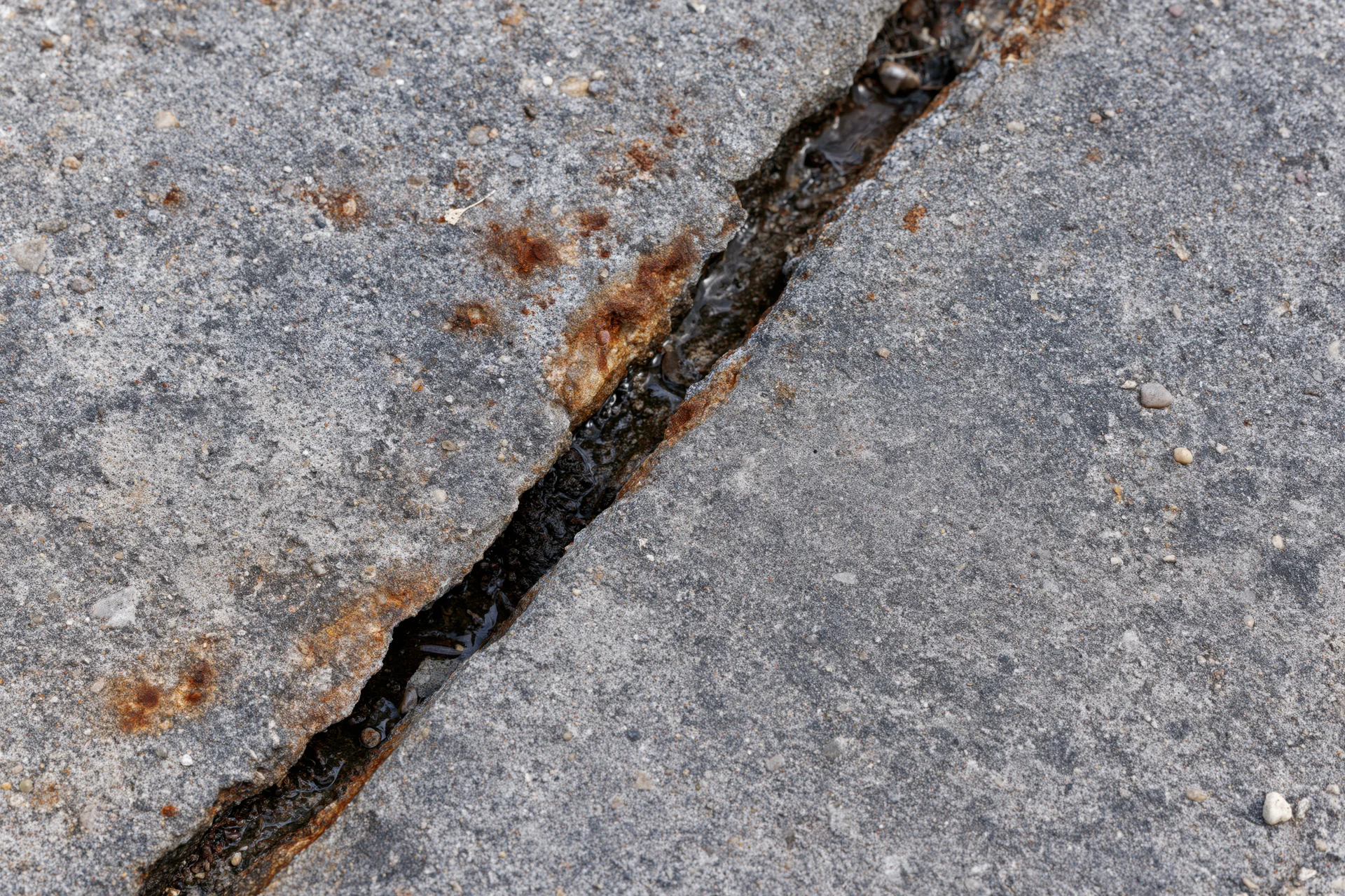 Diagonal crack in gray concrete pavement with rust and water accumulation