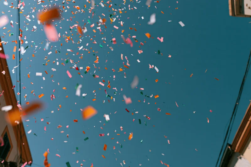 Colorful confetti floating upward in a clear blue sky above a narrow urban street
