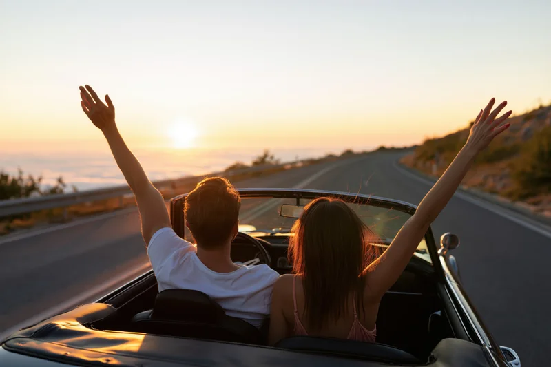 Couple driving a classic convertible along a winding coastal road at sunset