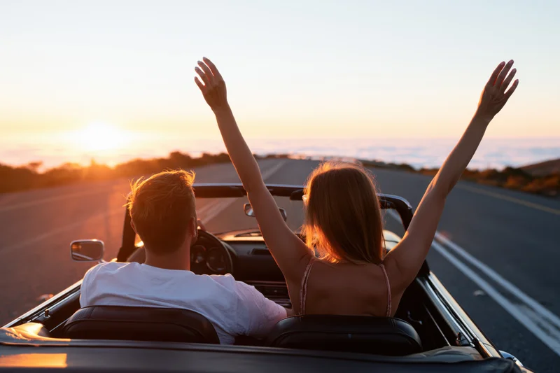 Couple driving a classic convertible on a winding mountain road at sunset