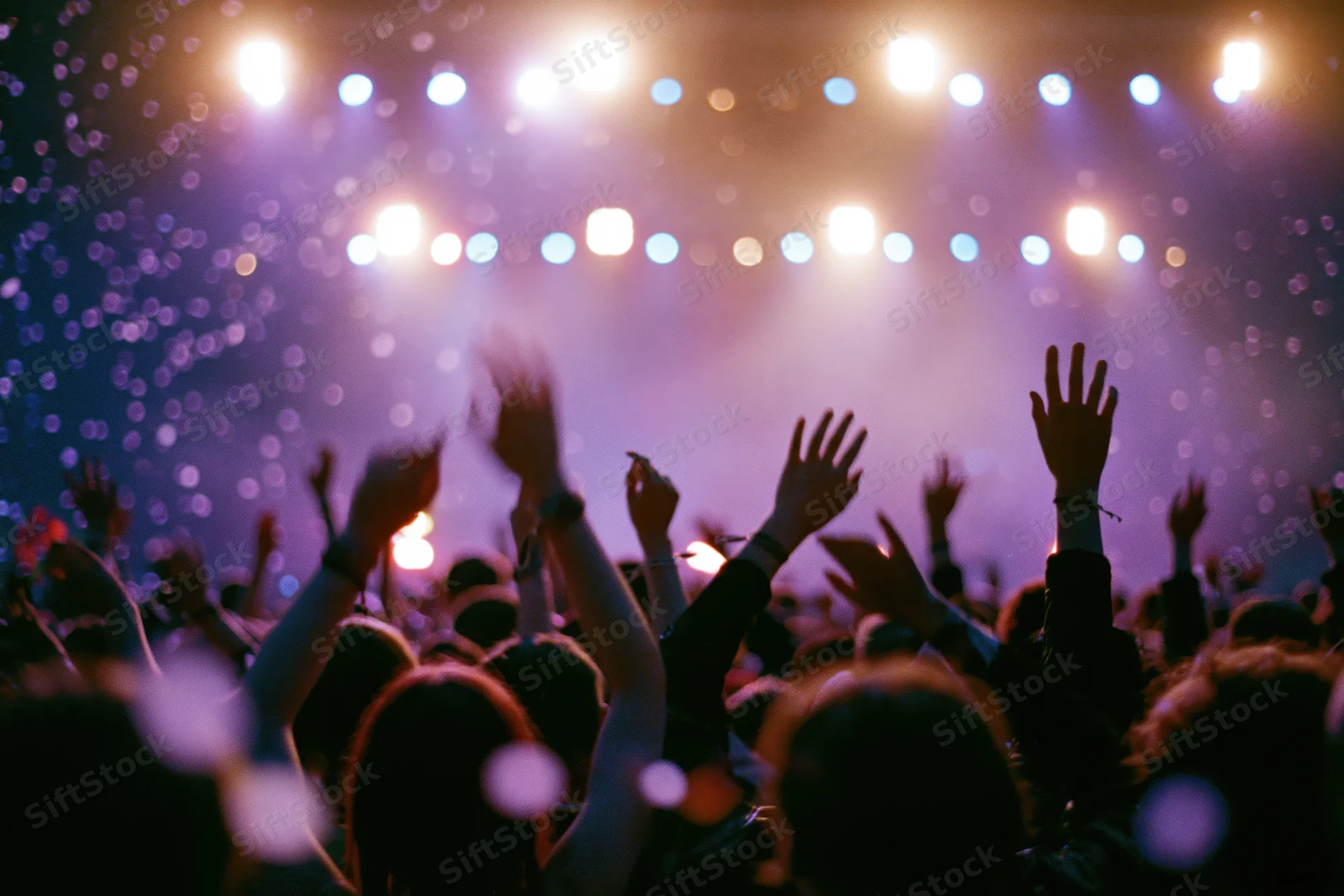 Crowd of concertgoers with arms raised silhouetted against stage lights