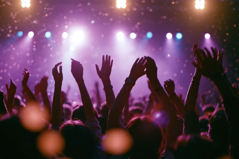 Crowd of concert-goers with arms raised silhouetted against stage lights