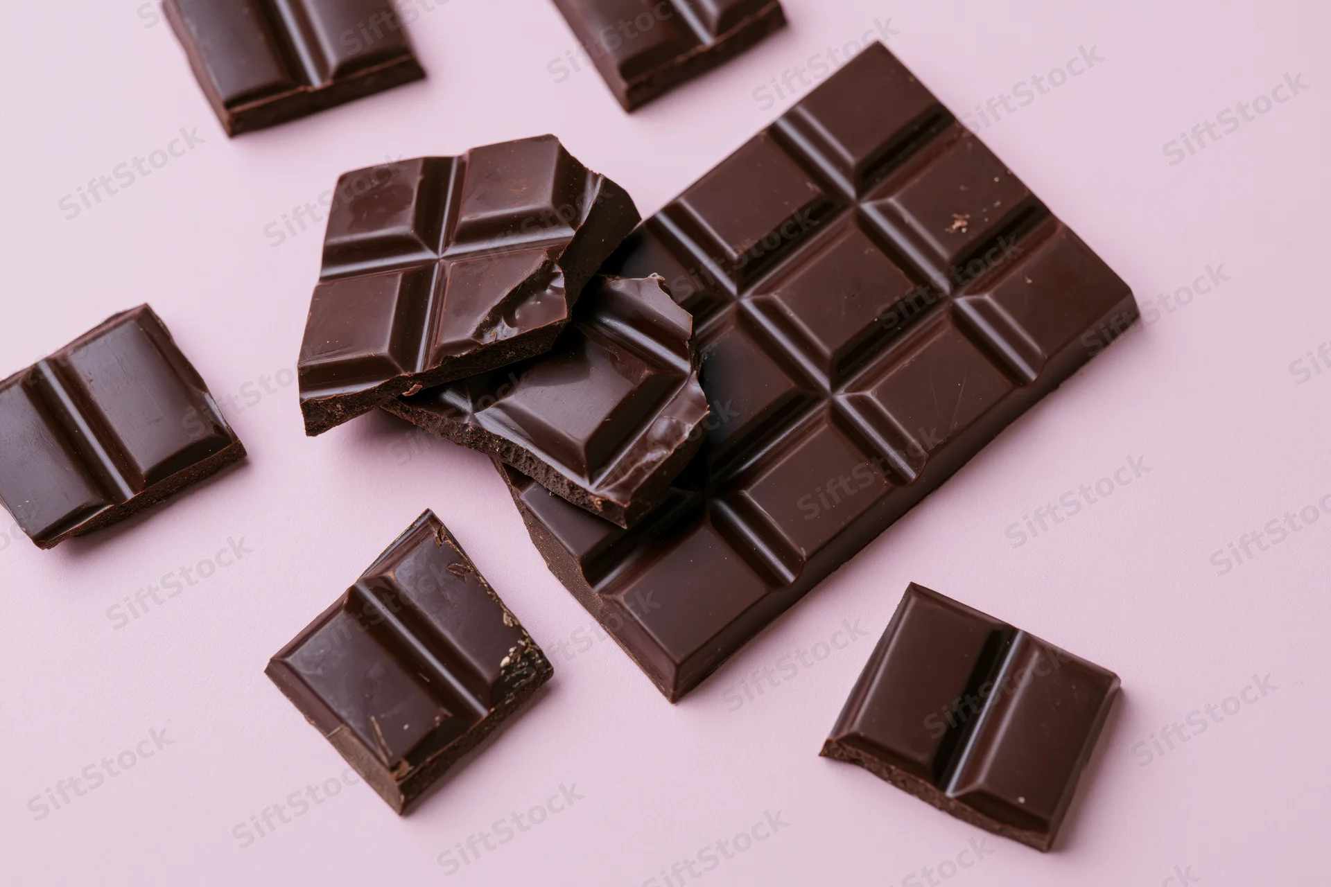 Broken dark chocolate bar pieces scattered on pink surface in flat lay arrangement