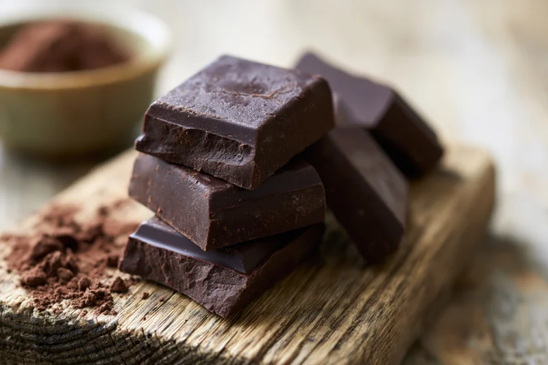 Thick dark chocolate chunks stacked on a wooden cutting board with cocoa powder nearby