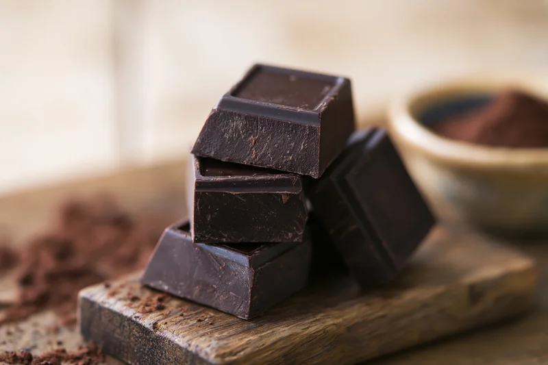 Thick dark chocolate chunks stacked on a wooden cutting board with cocoa powder nearby