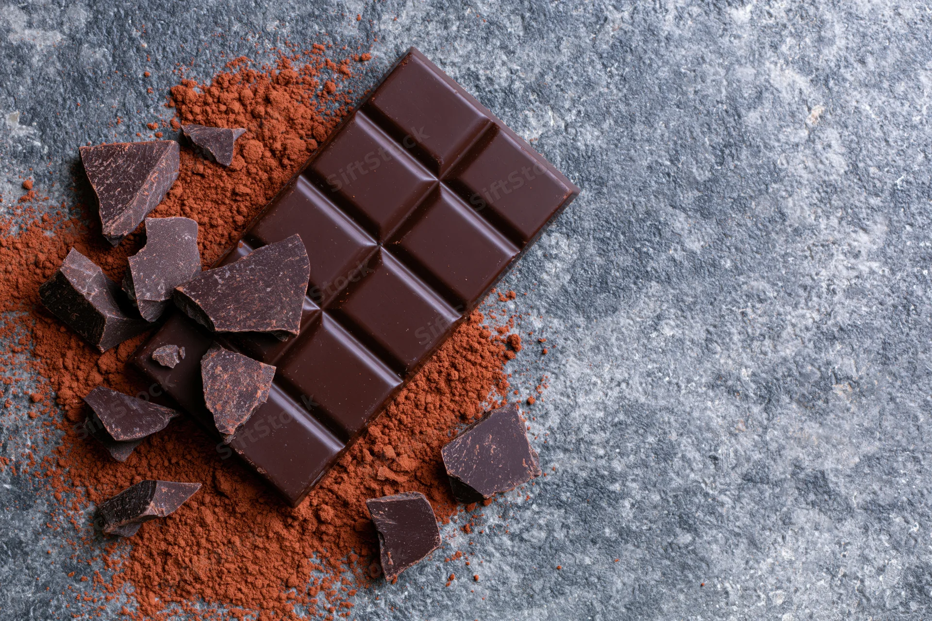 Dark chocolate bar with broken pieces and cocoa powder on a gray stone surface