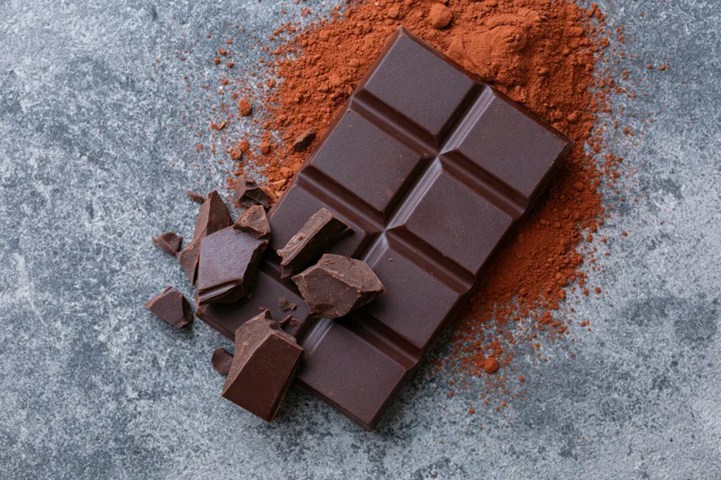 Dark chocolate bar with broken pieces and cocoa powder on a gray stone surface