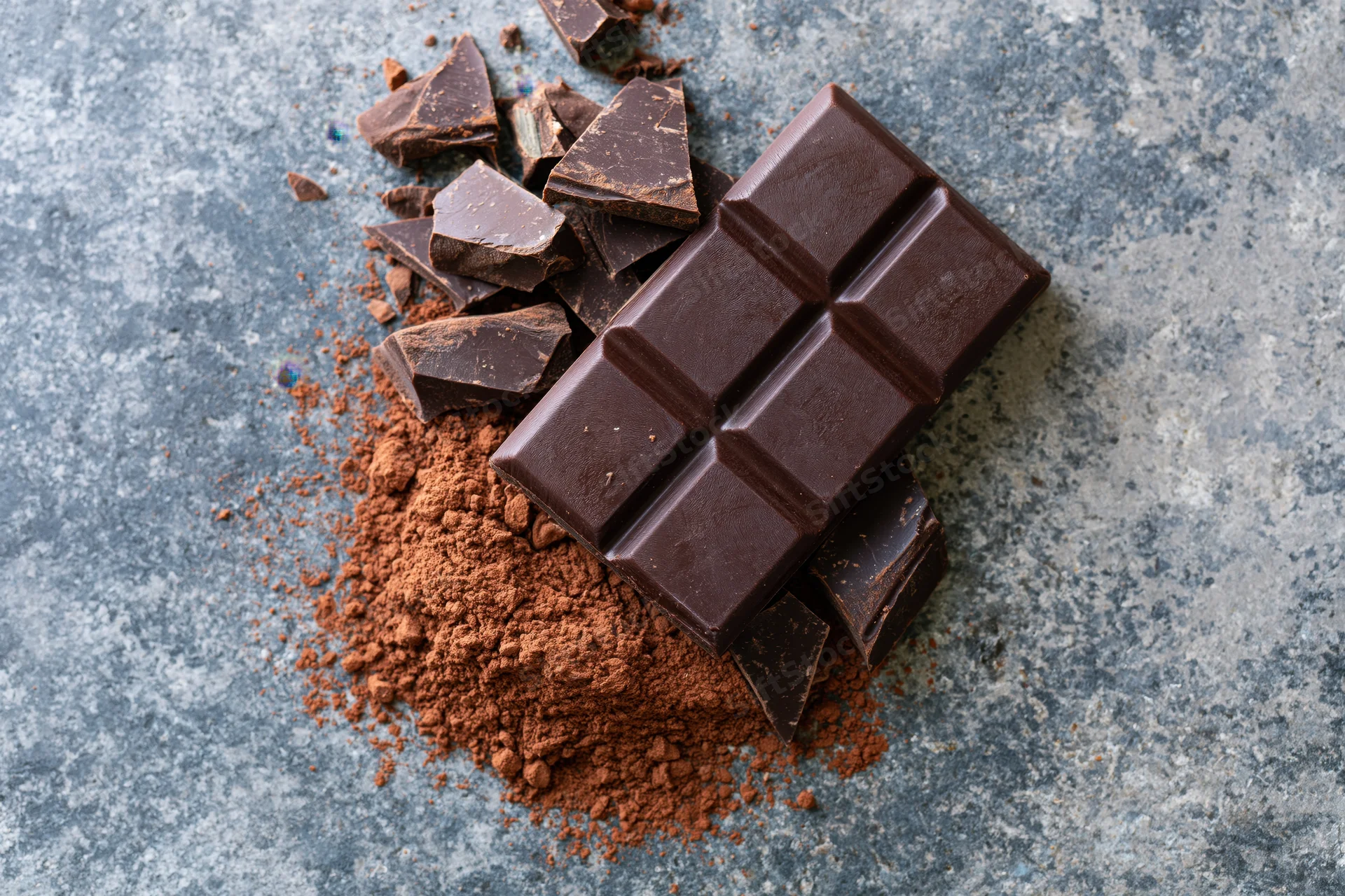 Dark chocolate bar with broken pieces and cocoa powder scattered on a gray stone surface