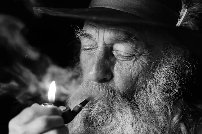 Elderly man with long gray beard lighting a pipe while wearing a hat with feather, close-up portrait in black and white