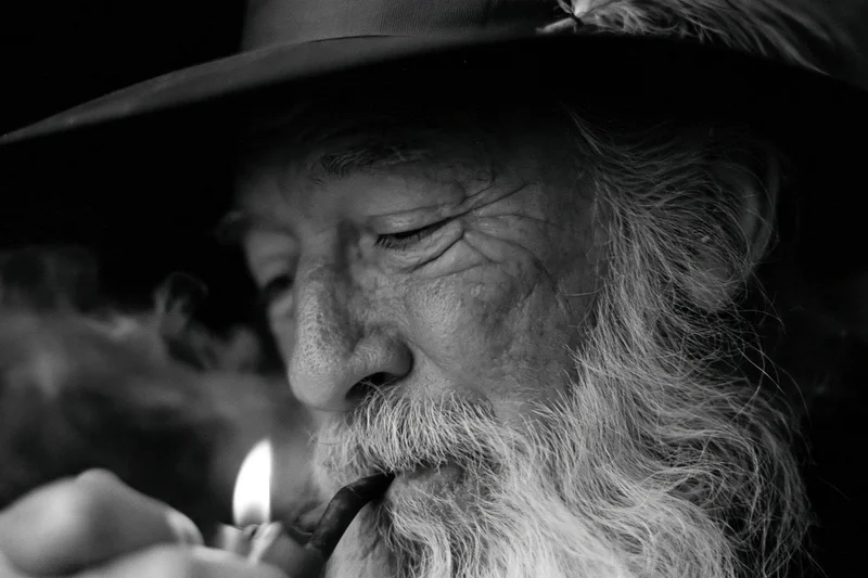 Elderly man with long silver beard lighting a pipe in close-up black and white portrait