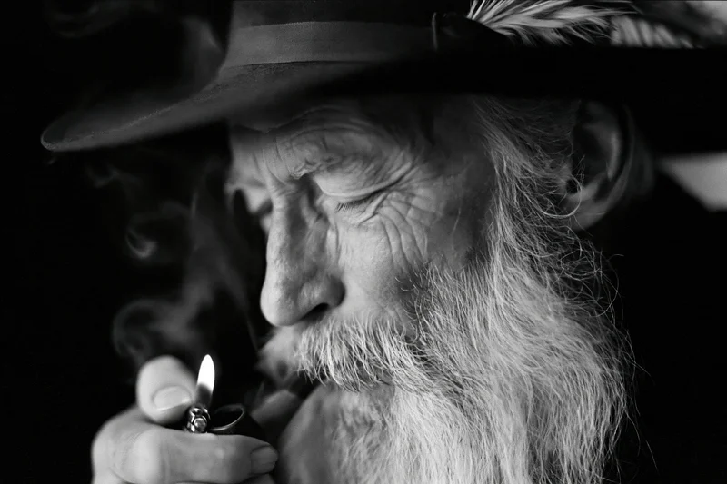 Elderly man with long silver beard lighting a pipe in profile, wearing a hat with feather, black and white closeup