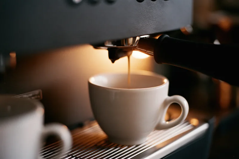 Espresso machine pouring dark coffee into a white ceramic cup on a drip tray in a cafe