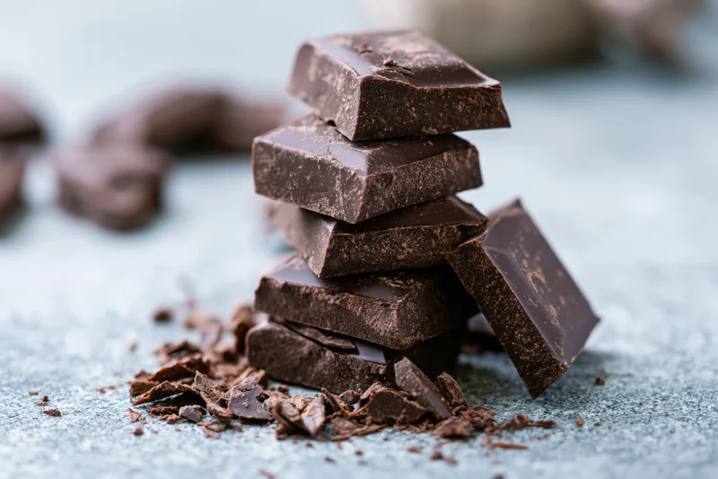 Broken dark chocolate squares stacked unevenly on a gray stone surface with crumbs scattered around