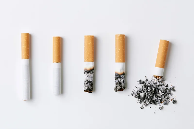 Five cigarettes in a row showing progressive stages of burning from unlit to fully extinguished and crushed on a white surface