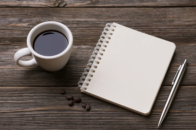 White ceramic mug with black coffee, spiral notebook and pen arranged on dark wooden table