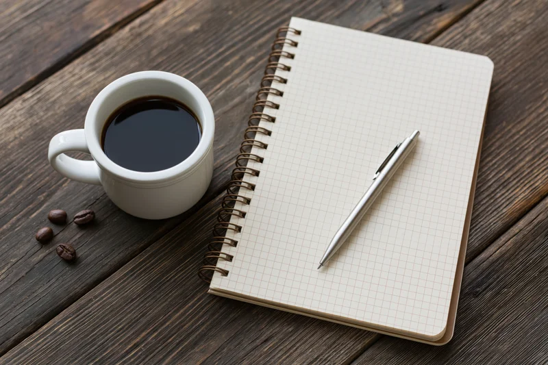 White ceramic mug with black coffee beside a spiral notebook and pen on a wooden table