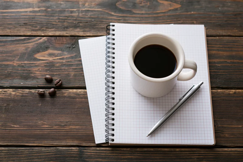 White ceramic mug with black coffee on a grid notebook beside a pen on dark wooden table