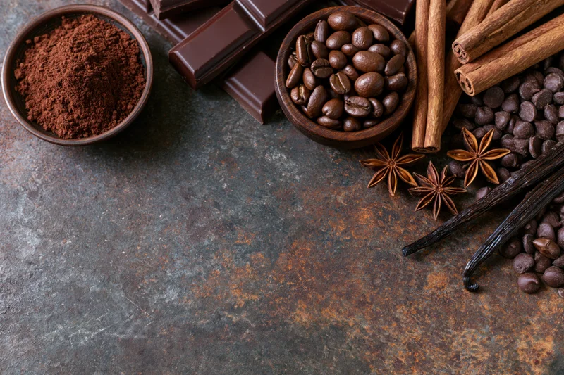 Chocolate bars, cocoa powder, coffee beans, cinnamon sticks, vanilla pods, and star anise arranged on a rusty metal surface in a flat lay composition