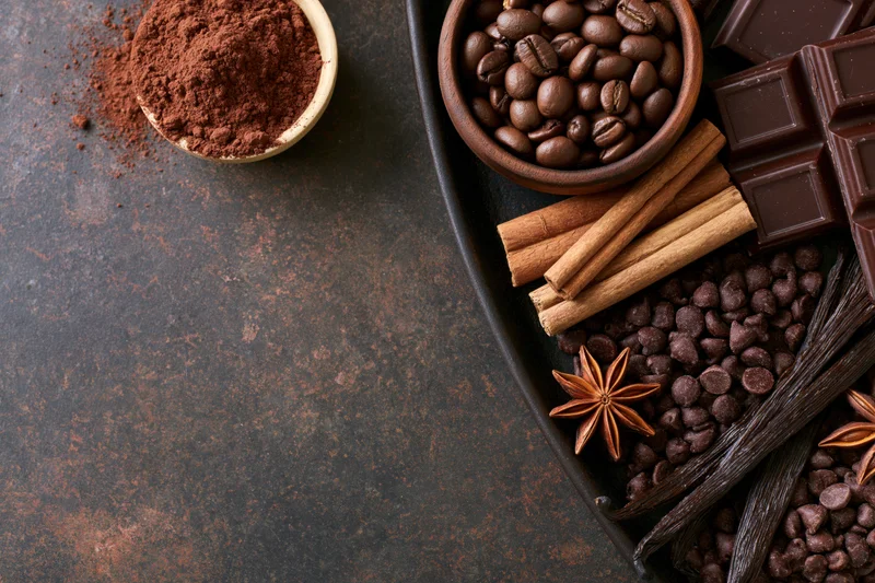 Chocolate bars, cocoa powder, coffee beans, cinnamon sticks, vanilla pods, and star anise arranged on a dark tray with copyspace on the left