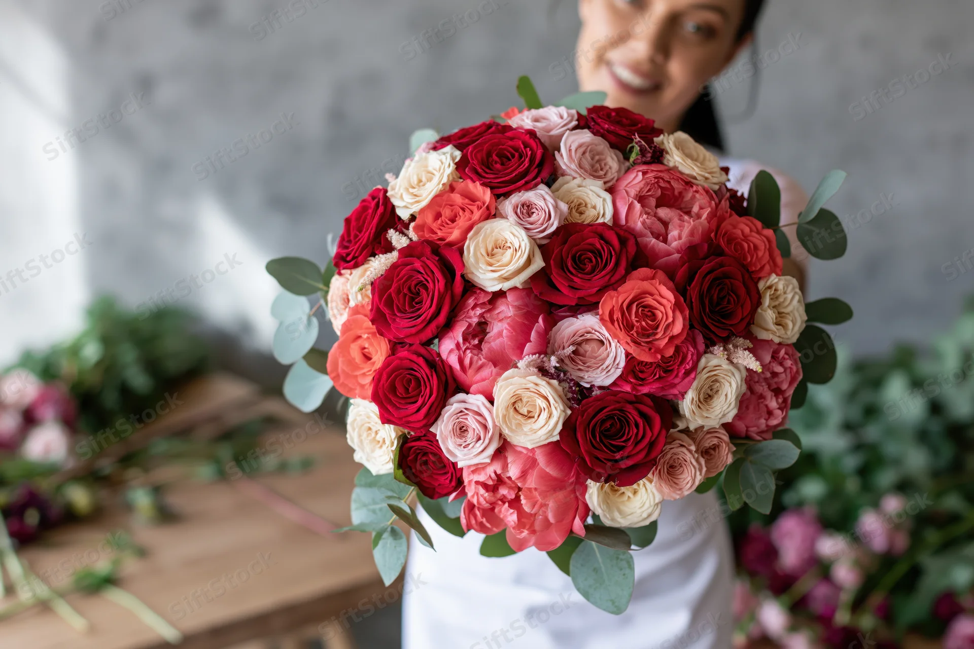 Florist holding large rounded bouquet of red roses, pink peonies, and eucalyptus in studio setting