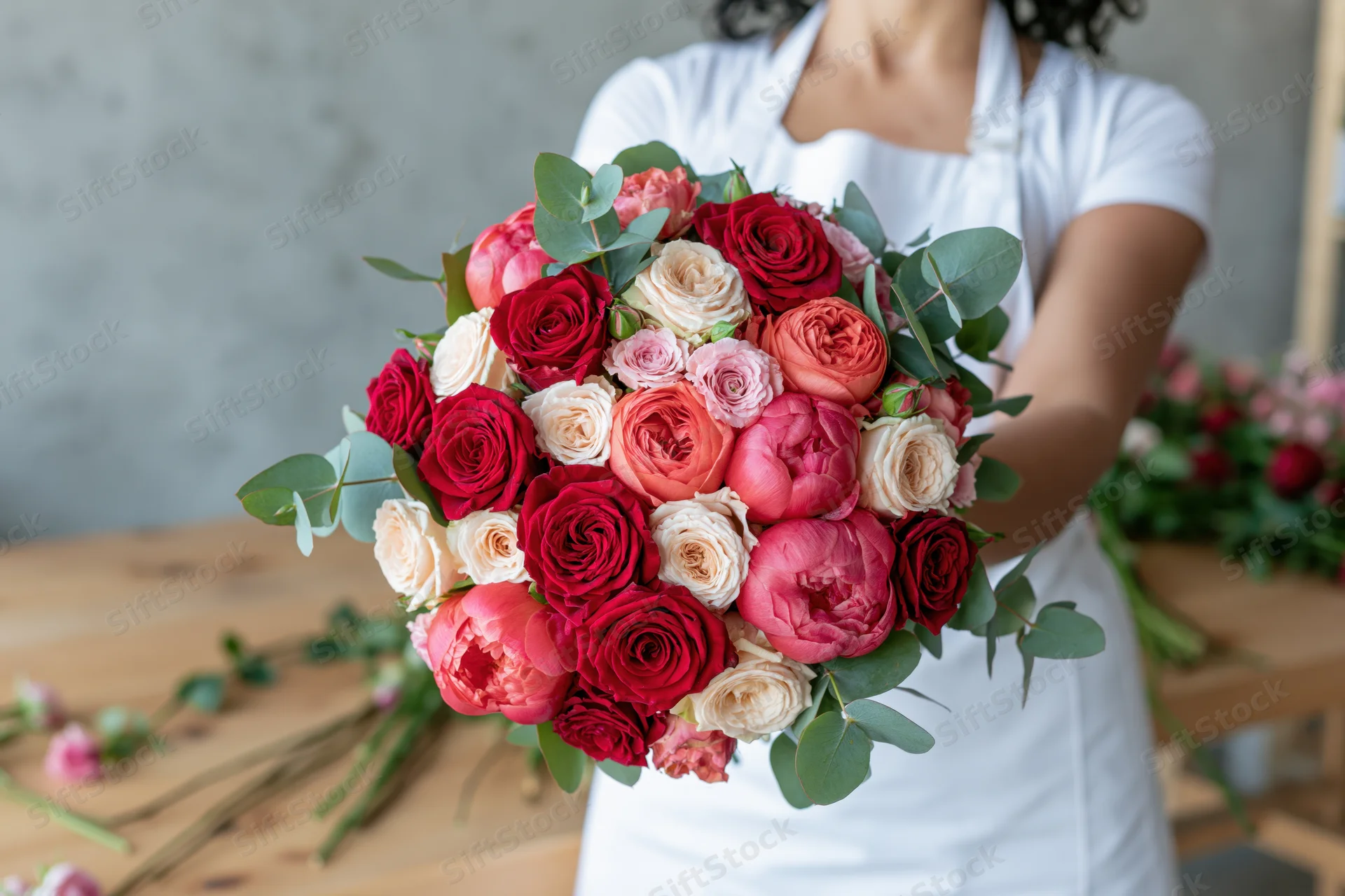 Florist holding a large rounded bouquet of red pink and coral roses with eucalyptus leaves on a wooden table