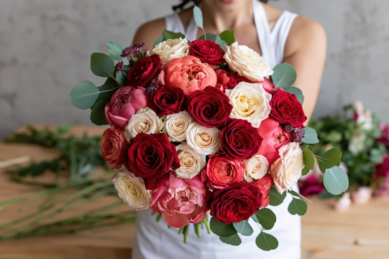 Woman holding large rounded bouquet of red roses and pink peonies with eucalyptus leaves in a workshop
